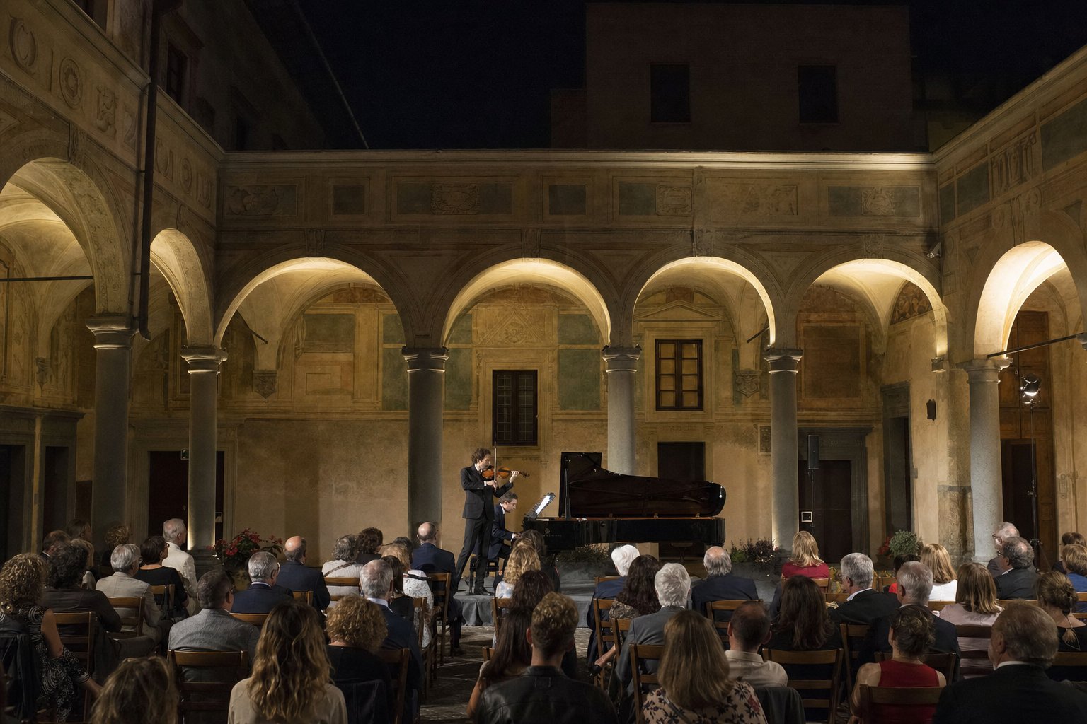 Chiostro medievale di San Marco a Tarquinia di sera con concerto in corso, colonne e archi illuminati, pubblico seduto ad ascoltare.