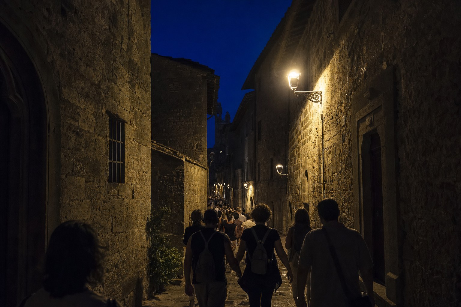 Vicolo medievale di Viterbo di notte con lampioni bassi, pietra tufacea illuminata e un gruppo di persone in cammino silenzioso.