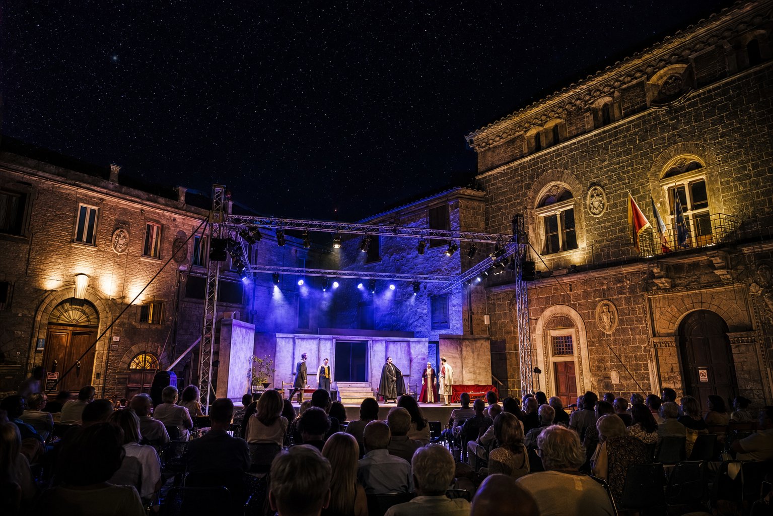 Palco teatrale in Piazza Umberto I a Vetralla di sera con attori in scena, palazzi storici illuminati e pubblico seduto ad ascoltare.